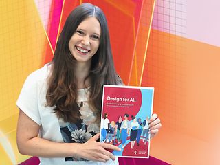 Annika Theiß hält den „Design für Alle Guide“ in der Hand. Sie steht vor einem bunten Design-Hintergrund.