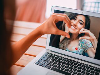 Two women, one in front and one on the computer screen, forming a heart together with teir hands.