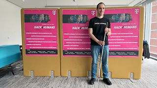 Mann mit einem Blindenstock vor Plakaten mit der Aufschrift „Hack4Humans 2024“