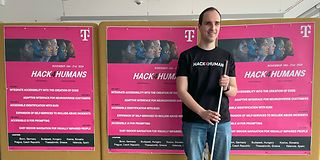 Mann mit einem Blindenstock vor Plakaten mit der Aufschrift „Hack4Humans 2024“