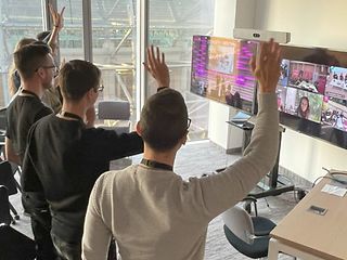 A team waves into a camera in front of a screen.