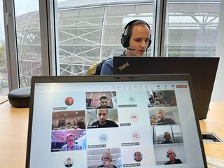 Laptop monitor with participants in a video conference. Behind it a man with a headset on a laptop.