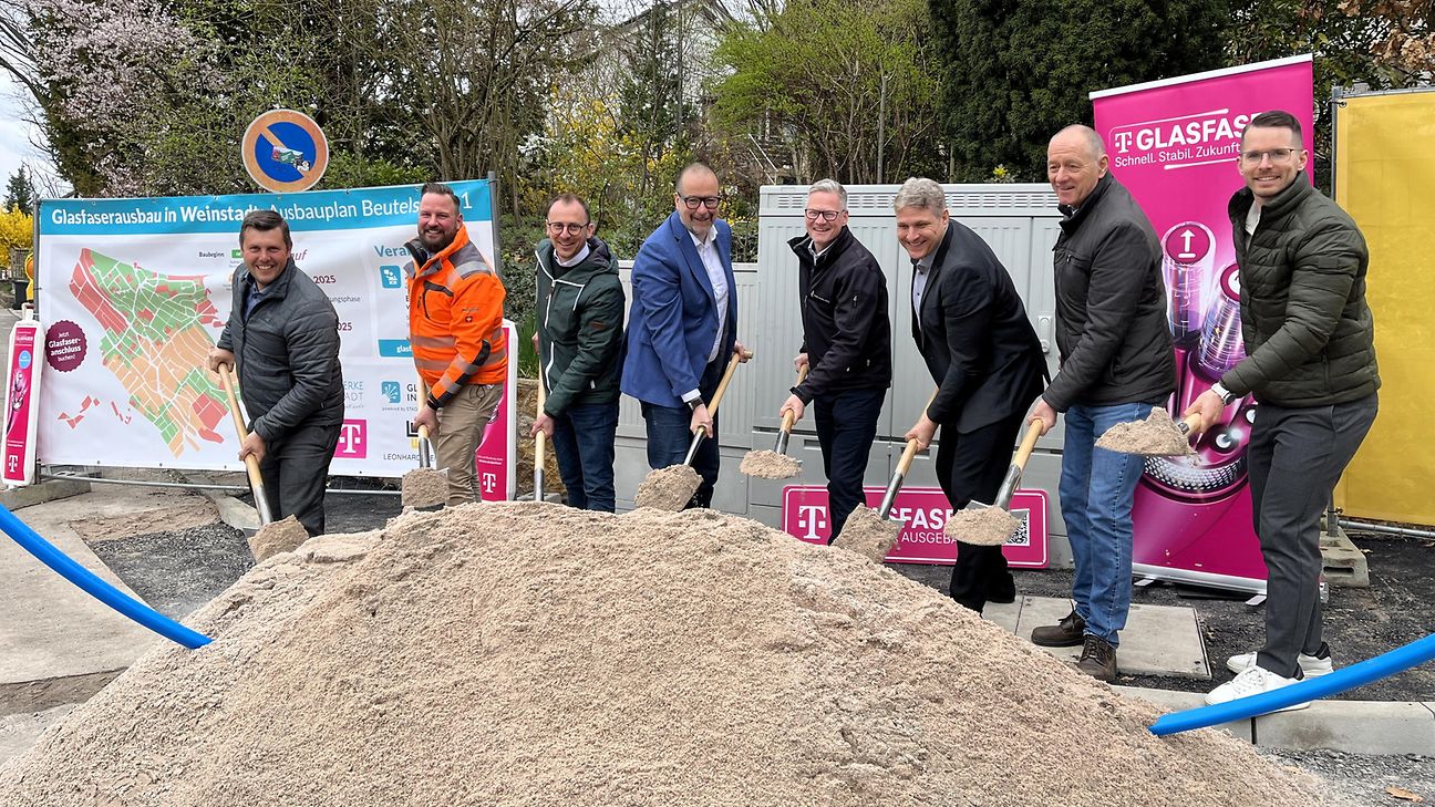 Personen stehen hinter einem großen Sandhaufen und schaufeln gemeinsam für den symbolischen Spatenstich. 