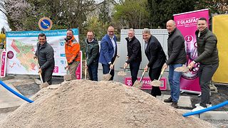Personen stehen hinter einem großen Sandhaufen und schaufeln gemeinsam für den symbolischen Spatenstich. 