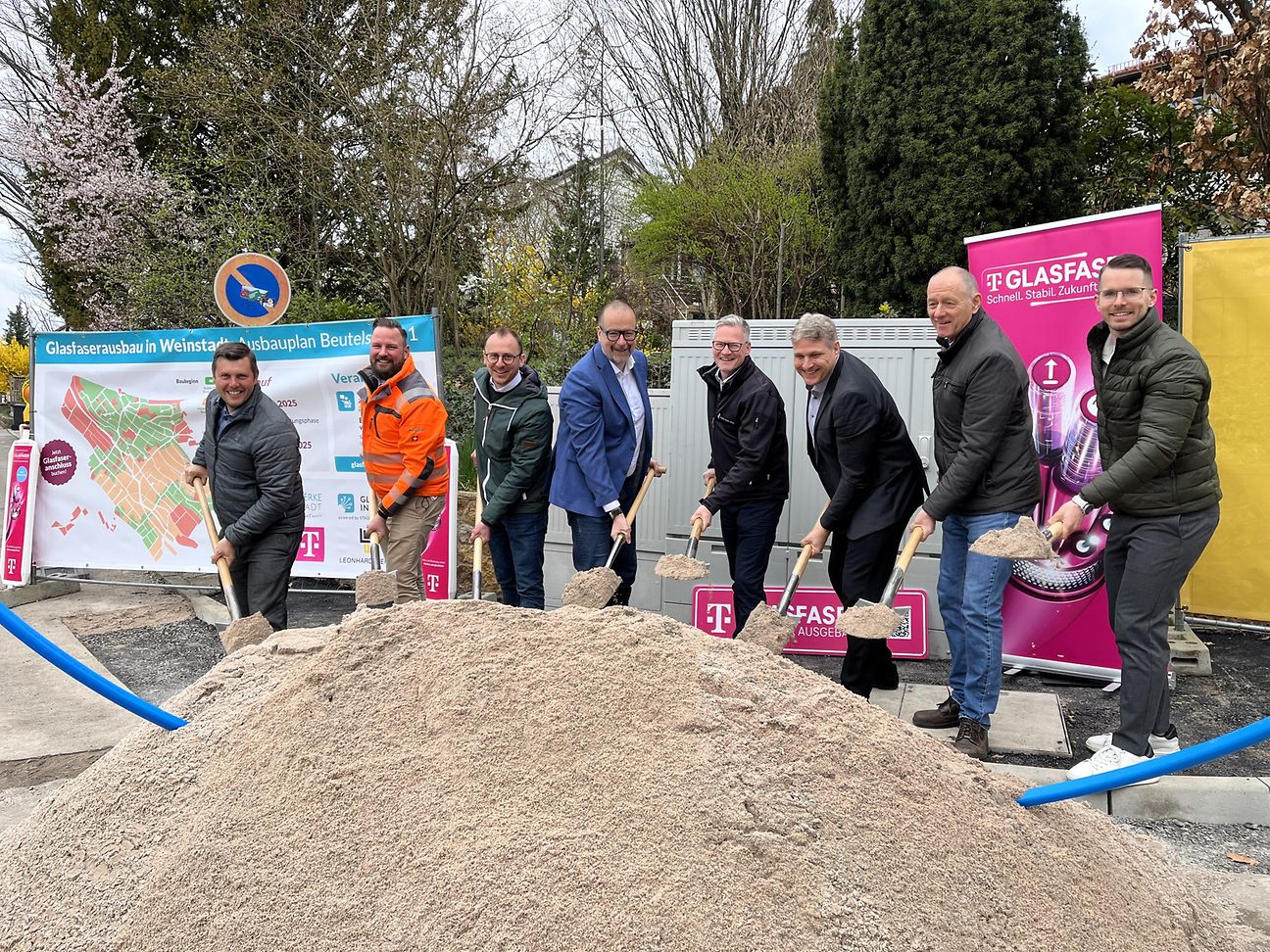 Personen stehen hinter einem großen Sandhaufen und schaufeln gemeinsam für den symbolischen Spatenstich. 