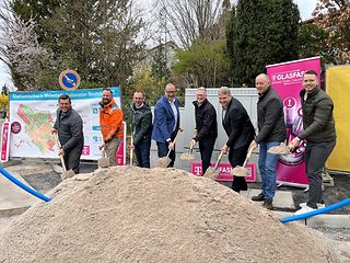 Personen stehen hinter einem großen Sandhaufen und schaufeln gemeinsam für den symbolischen Spatenstich. 