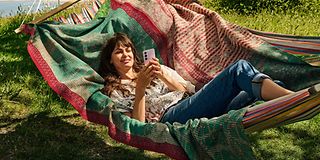 Junge Frau liegt in einer Hängematte und führt mit ihrem Smartphone ein Videotelefonat. 