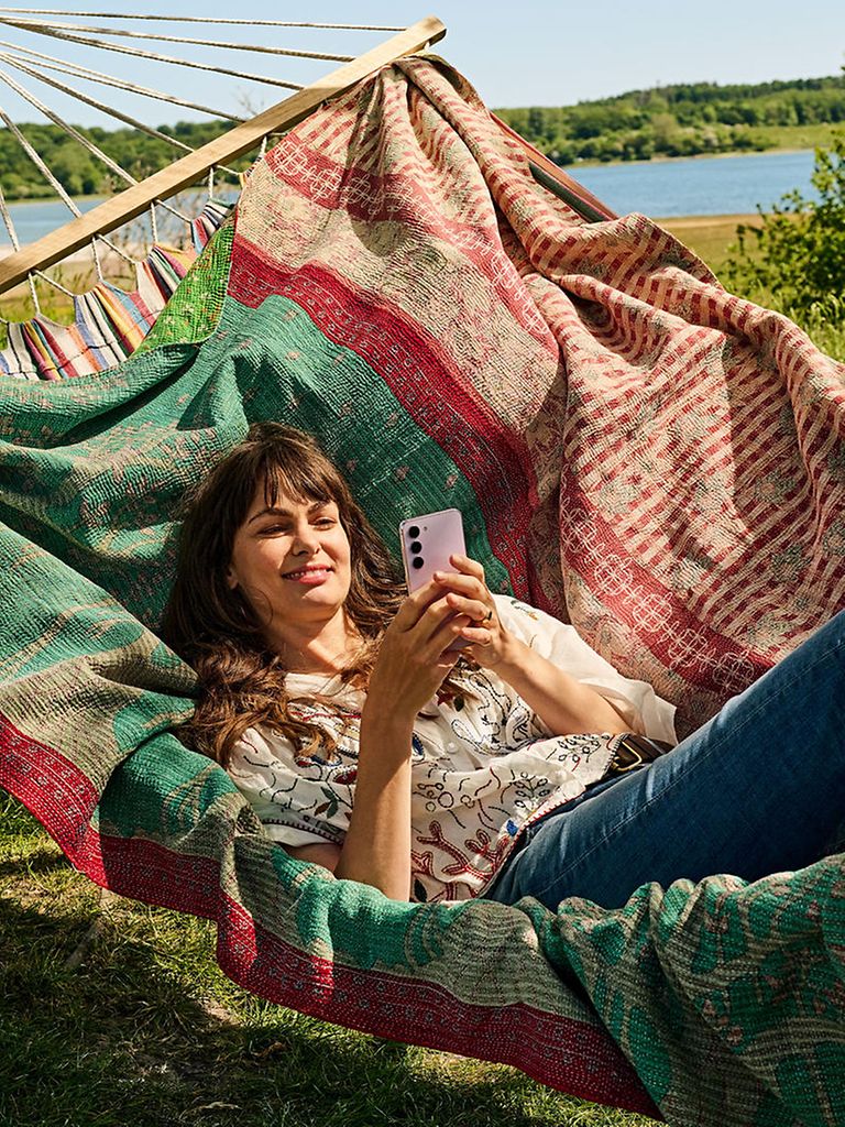 Junge Frau liegt in einer Hängematte und führt mit ihrem Smartphone ein Videotelefonat. 