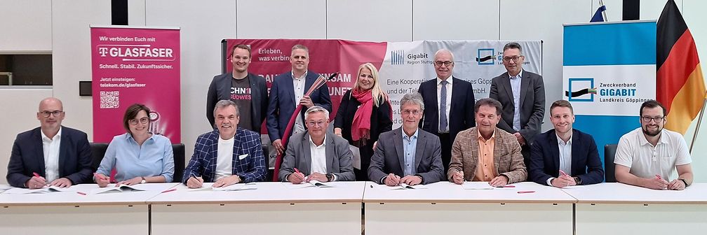 Vertreter der Kommunen sitzen mit Unterschriftenmappen an einer Tischreihe, die übrigen Personen stehen dahinter.