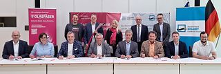 Vertreter der Kommunen sitzen mit Unterschriftenmappen an einer Tischreihe, die übrigen Personen stehen dahinter.