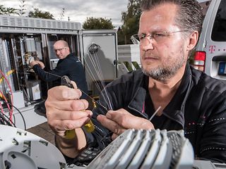 Zwei Techniker arbeiten am Ausbau des Breitbandnetzes. Ein Techniker kürzt mit einer Zange einzelne Glasfasern ein. 