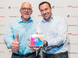 Michael Schuster and Ronald Schulz laughing as they hold the award up to the camera – a colorful Plexiglas cube.