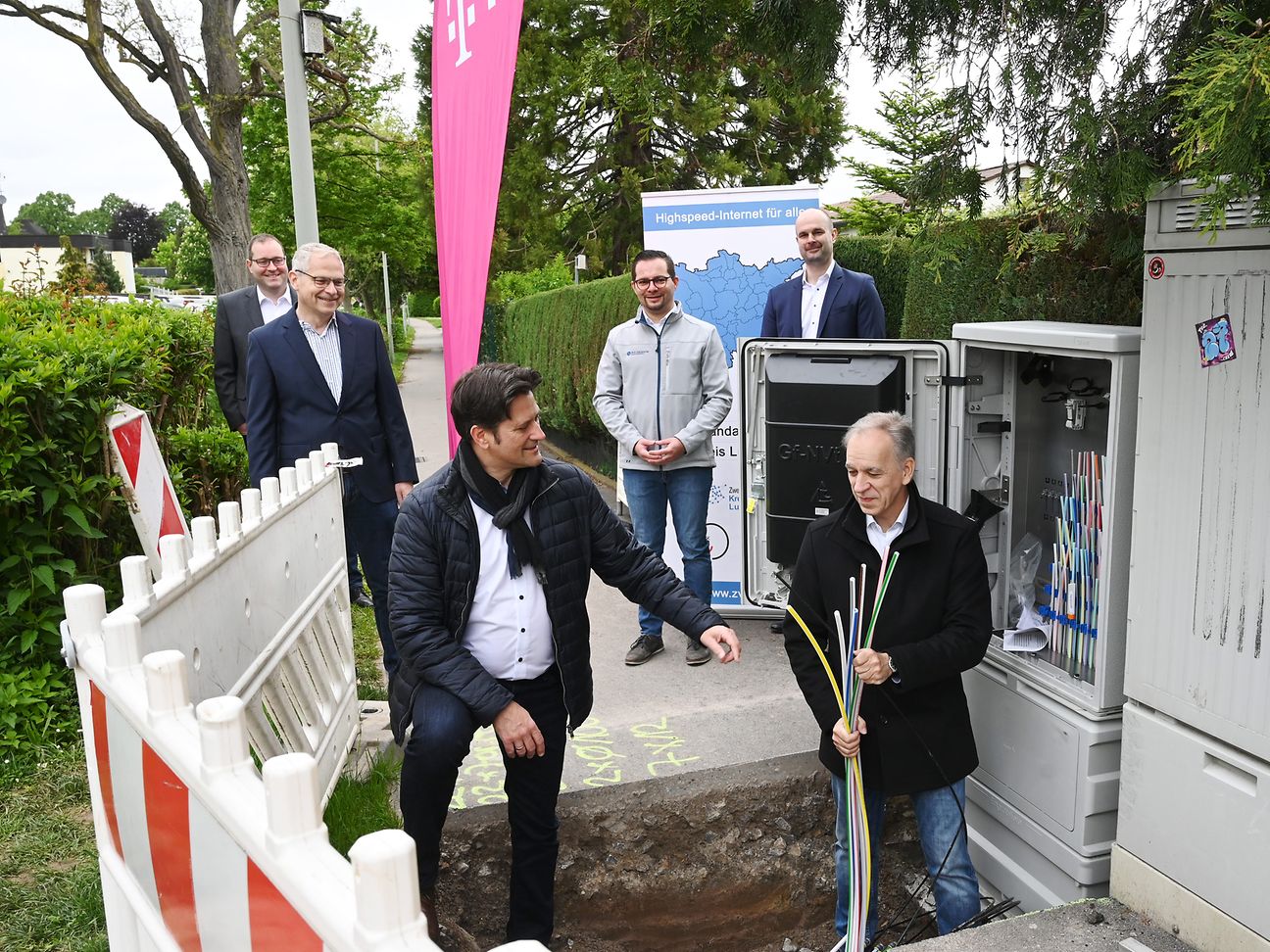 Volker Ackermann und Dirk Schaible stehen in der Baugrube vor einem Verteilerkasten, drum herum weiteren Personen. 