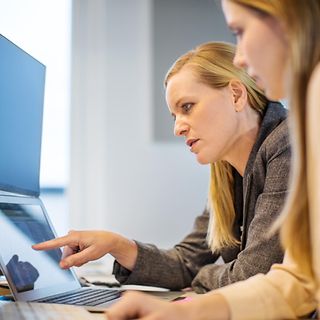 Eine Frau erklärt einer anderen etwas an einem Laptop in einem Büro