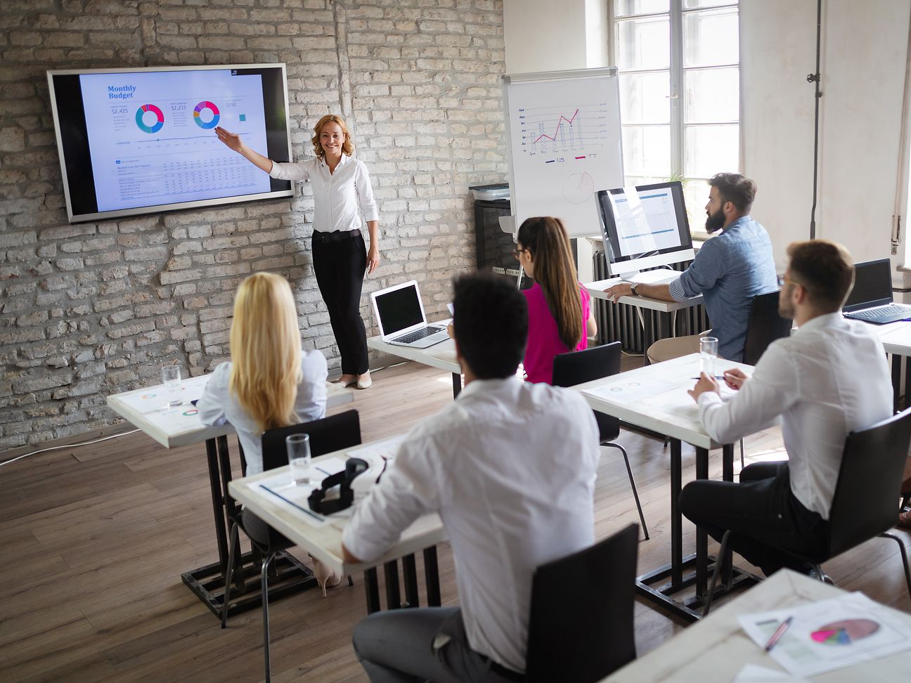 Lehrerin steht vor digitalem Board, Gruppe von Schüler*innen sitzt am Schreibtisch mit Laptops..