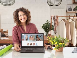 Frau sitzt am Schreibtisch in einem Geschäft und zeigt den Bildschirm ihres Laptops. Darauf ist ihre Webseite zusehen.